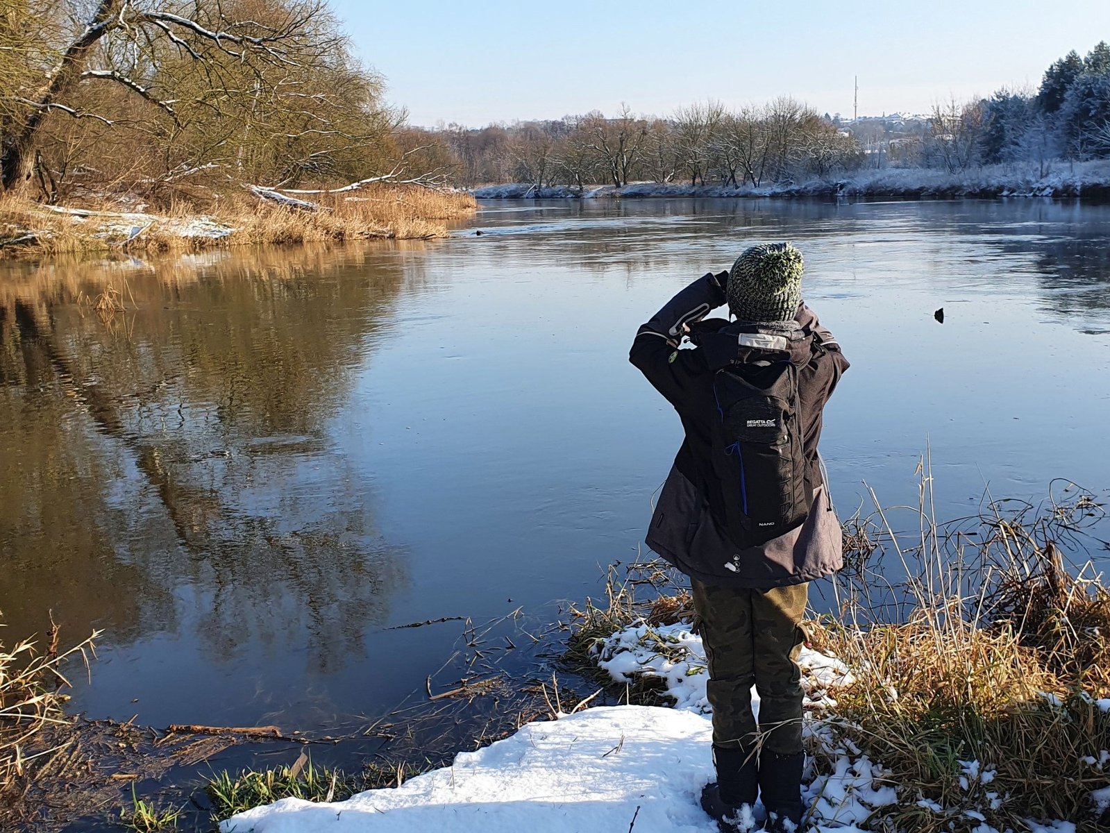 Osoba stoi nad brzegiem rzeki w zimowej aurze, nad jej brzegami nachylają się drzewa.