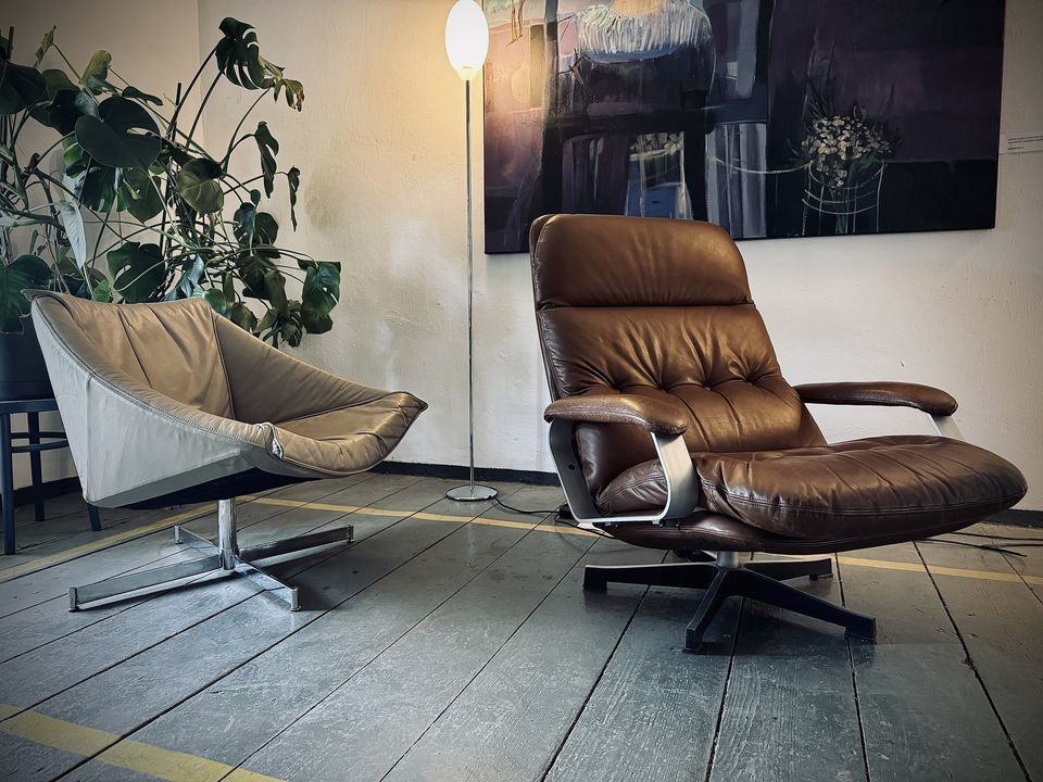 Zdjęcie przedstawia przytulny kącik wewnętrzny, który można znaleźć w domu, biurze lub salonie. Znajdują się w nim dwa różne rodzaje krzeseł ustawionych naprzeciwko siebie, tworząc przestrzeń do rozmów. Po lewej stronie znajduje się krzesło w nowoczesnym stylu z jasnoszarą tapicerką z tkaniny i minimalistyczną metalową ramą. Po prawej stronie znajduje się pluszowa skórzana leżanka w bogatym brązowym kolorze z drewnianymi akcentami na podłokietnikach, co sugeruje wygodę i relaks.