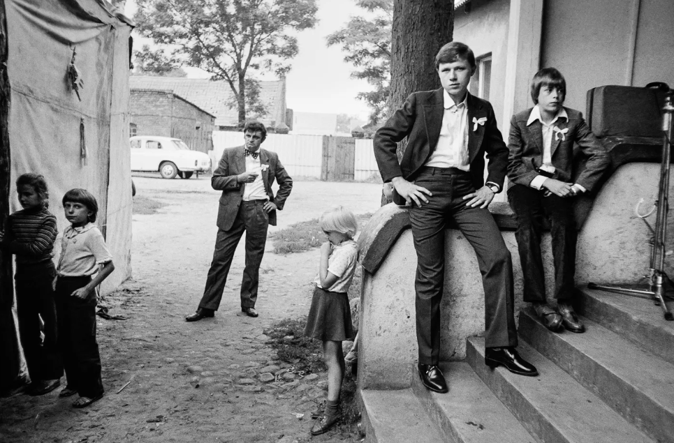 Czarnobiałe zdjęcie grupy młodych mężczyzn i dzieci. Mężczyźni ubrani są w retrro garnitury. Dzieci ubrane są w ciemne spodnie i jasne koszulki. Pozują na schodach pod budynkiem.