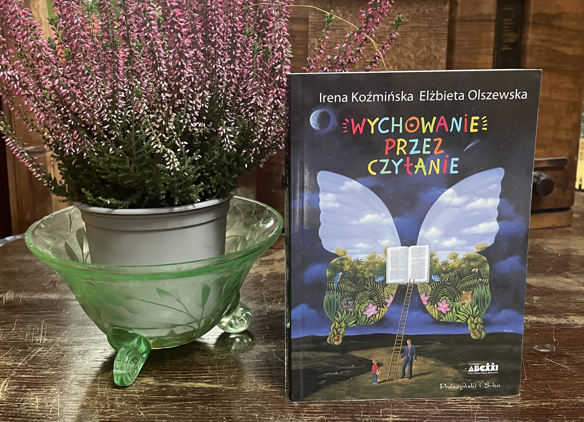 Zdjęcie, obok doniczki z wrzosami stoi książka, pt."Wychowanie przez czytanie", Irena Koźmińska, Elżbieta Olszewska. Na okładce znajduje się otwarta książka, z której wyrastają ogromne skrzydła motyla. Do ksiązki prowadzi drabina, przed którą stoi dziecko i dorosły mężczyzna..