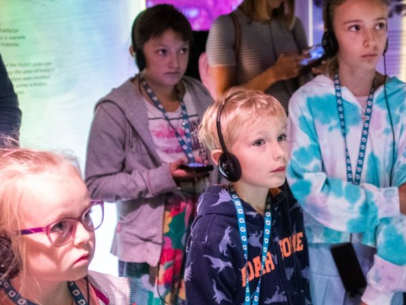 Grupa dzieci stoi, słuchając czegoś przez słuchawki.