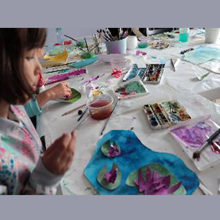 Stół z farbami i przyborami malarskimi, po lewej stronie dziewczynka z pędzelkiem