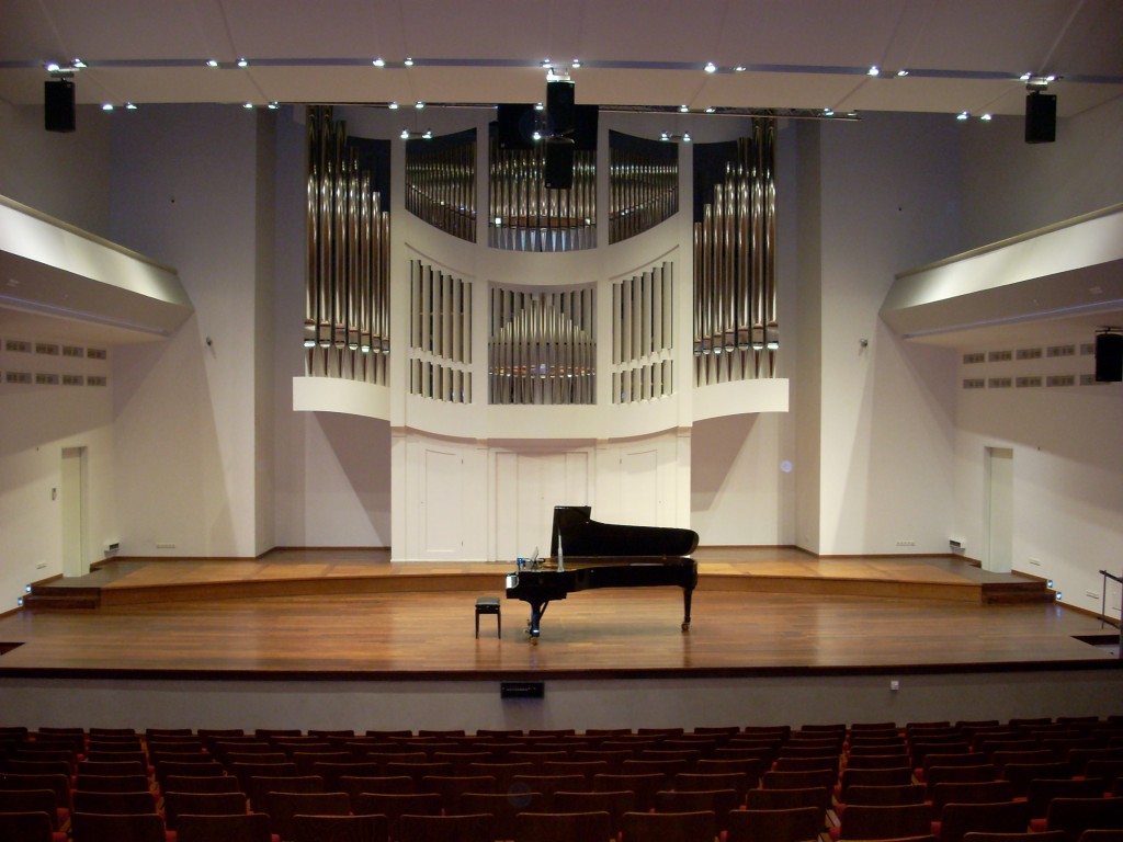 Zdjęcie przedstawia scenę auli Akademii Muzycznej w Poznaniu. Na środku sceny ustawiony jest fortepian. W tle widoczne piszczałki organów.