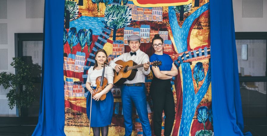 Zdjęcie trzyosobowego zespołu muzycznego (gitara, skrzypce i wokal). W tle namalowany kolorowy plan miasta.