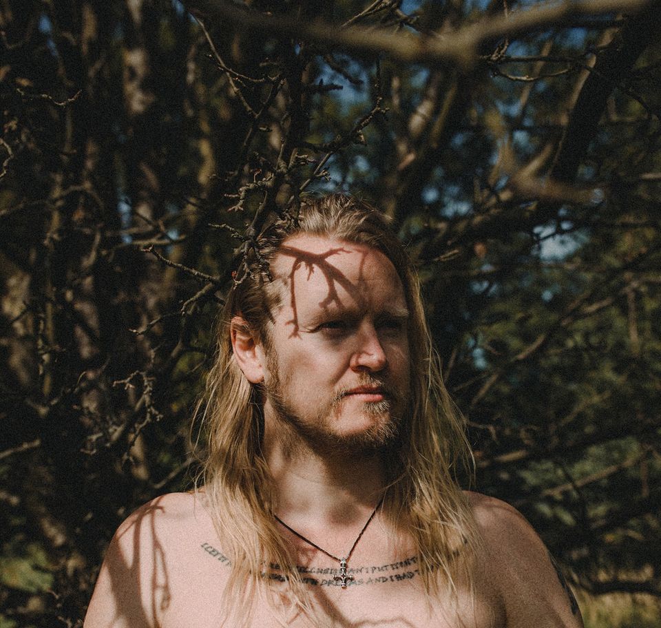 zdjęcie wykonawcy. Długowłosy mężczyzna z zarostem na tle bezlistnego drzewa.