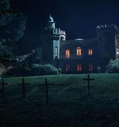 Kadr z filmu "Gniazdo zła" przedstawia gotycką posiadłość, stojącą na pagórku. Na zboczu pagórka krzyże. Jest ciemno i mroczno.