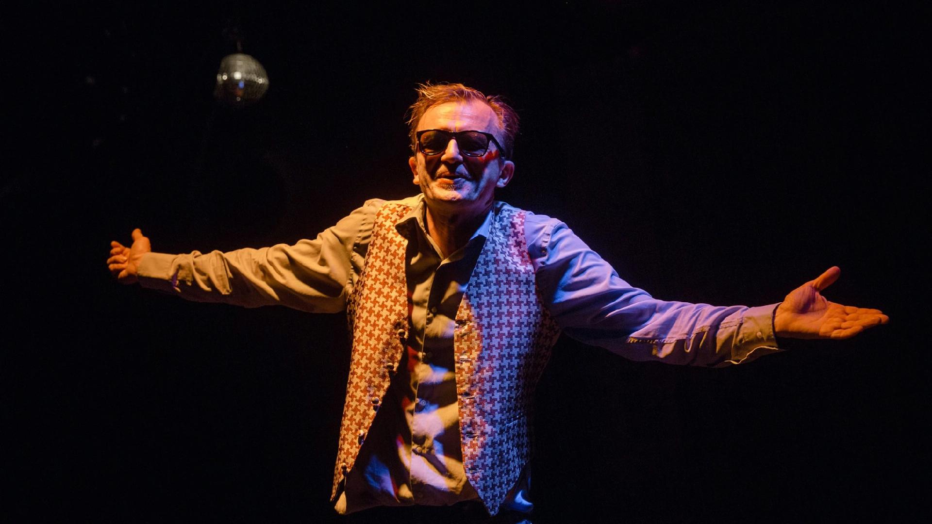 Na zdjęciu na czarnym tle, aktor w ciemnych okularach z rozłożonymi ramionami.