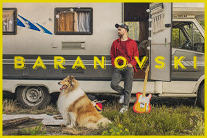 Na zdjęciu mężczyzna opierający się o przyczepę campingową. W otwartych drzwiach przyczepy stoi gitara. Przed przyczepą leży pies.