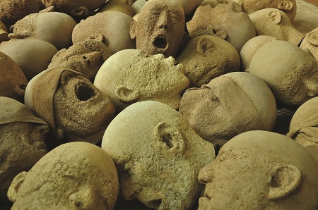 Fotografia z wystawy Andrzeja Okińczyca "W Imię Boga Jedynego" przedstawia liczne rozrzucone, chaotycznie kamienne głowy z twarzami zastygłymi w krzyku.