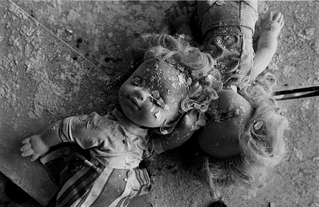 Czarno-białe zdjęcie przysypanych fragmentami pokruszonego tynku dwóch lalek, leżących na zniszczonym blacie stołu.