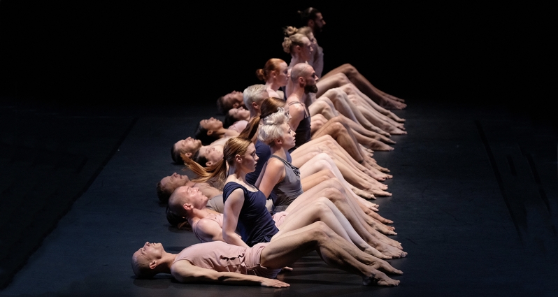 Grupa tancerzy leżących na scenie jeden za drugim. Niektórzy z nich są w trakcie podnoszenia się.