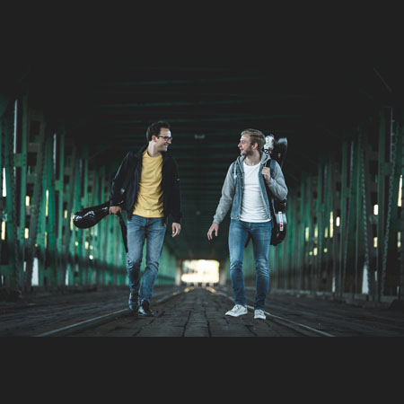 Dwaj muzycy idący po moście z instrumentami. Są zwróceni twarzami do siebie, uśmiechają się.