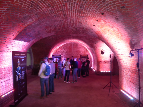 Grupa ludzi w podziemiach kaponiery, czytają plakaty rozstawione w pomieszczeniu.