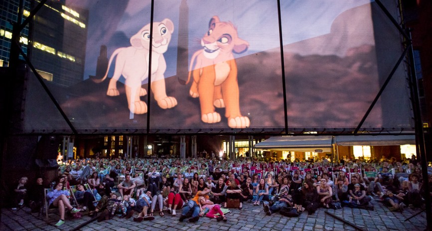 Grupa ludzi siedząca na ziemi pod ekranem z wyświetlonym filmem "Król lew".