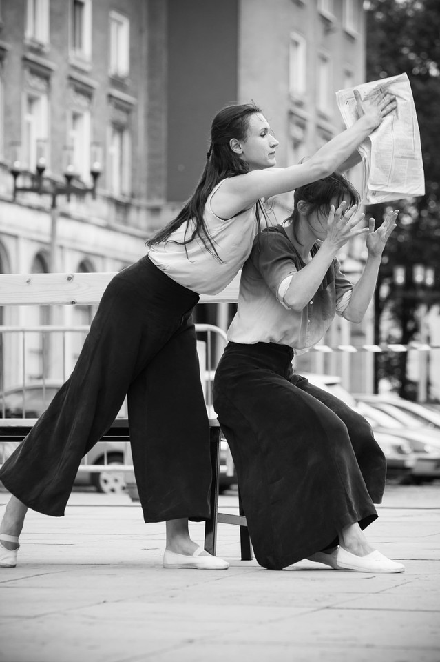 Zdjęcie czarno-białe. Dwie kobiety ujete w momencie wykonywania sceny. Jedna siedzi, a druga stoi nad nią i trzyma gazetę.