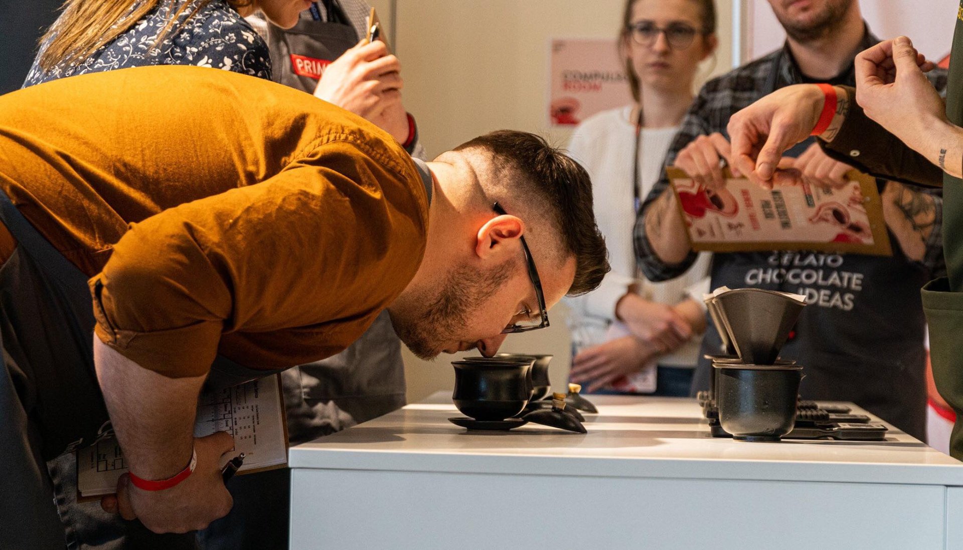 Zdjęcie przedstawiające mężczyznę schylonego i wąchającego napój w filiżance, obserwuje go grupa ludzi