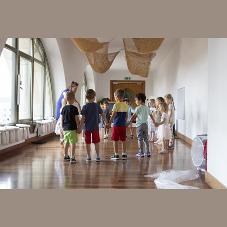 Na zdjęciu dzieci stoją na drewnianej podłodze w Bawialni w kole i trzymają się za ręce. Po lewej stronie widzimy prowadzącą zajęcia, a za nią duże okno w zielonej ramie.