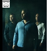 Zdjęcie przedstawia trzech muzyków w koszulach stojących na butelkowym tle.