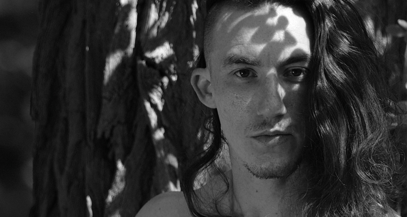 Czarno białe zdjęcie przedstawia twarz mężczyzny z lekkim zarostem i długimi włosami na tle drzewa.