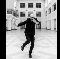 Czarno-biała fotografia. W centralnym punkcie Mariusz Lubomski w ruchu, w czarnym garniturze i okularach na nosie. Pozuje na tle dużego, jasnego korytarza, który z każdej strony jest otoczony oknami i portalami.