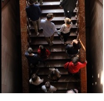 Grupa ludzi ukazana z perspektywy lotu ptaka wchodzi po schodach. Przed nimi okno; świetlają ich promienie zachodzącego słońca.