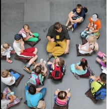 Grupa dzieci oraz osoba dorosła - przewodnik - siedzą na poduszkach rozłożonych na szarej kamiennej posadzce. Zdjęcie wykonane z perspektywy lotu ptaka.