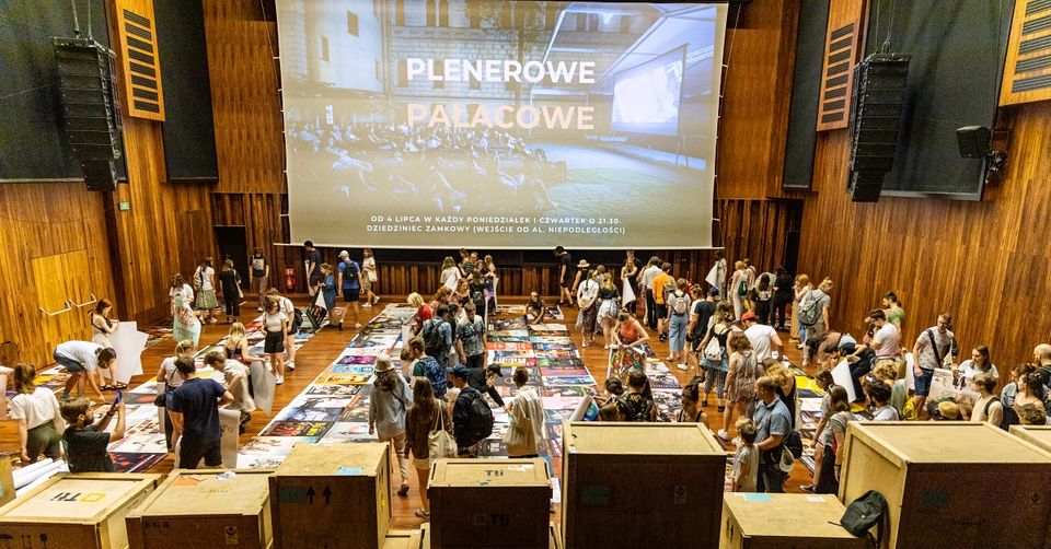 Plakaty rozłożone na ziemi zbierane i oglądane przez ludzi. Na ścianie wielki ekran z wyświetlonym napisem "Plenerowe Pałacowe". Sala jest cała drewniana (podłoga, ściany i sufit)