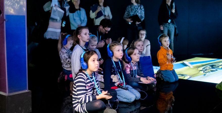Dzieci siedzące na podłodze w słuchawkach. Za nimi rodzice/opiekunowie. Wszyscy znajdują sie w sali multimedialnej. Na podłodze wywietlają się animacje.