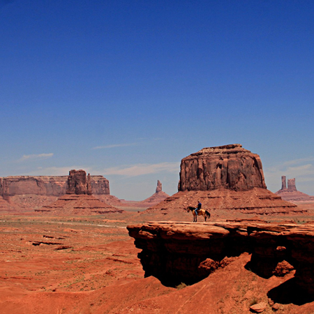 Kadr pochodzi z westernu Johna Forna "Nosiła żółtą wstążkę" i przedstawia panoramiczny widok na ikoniczną górę Oljato w Monument Valley w Colorado. Niebieskie niebo, czerwono-brunatne skały i piasek, na środku kadru widać kowboja na koniu, stojącego na małym wzniesieniu.