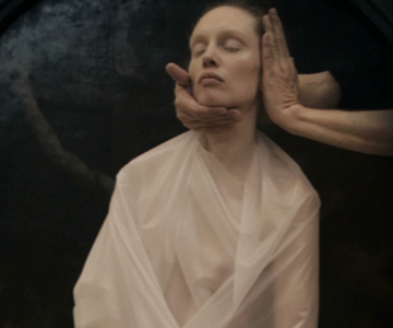 zdjęcie - siedząca kobieta w białej folii. Ma zamknięte oczy, a jej głowę trzymają dwie dłonie postaci stojącej obok.