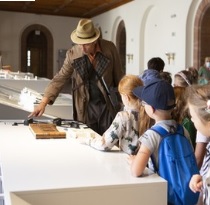 Zdjęcie detektywa w kapeluszu, a obok niego grupka dzieci - uczestników warsztatów.