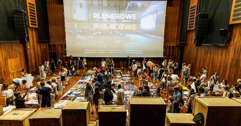 Drewniania sala z rozłożonymi plakatami na ziemi, a wśród nich ludzie wybierający je.