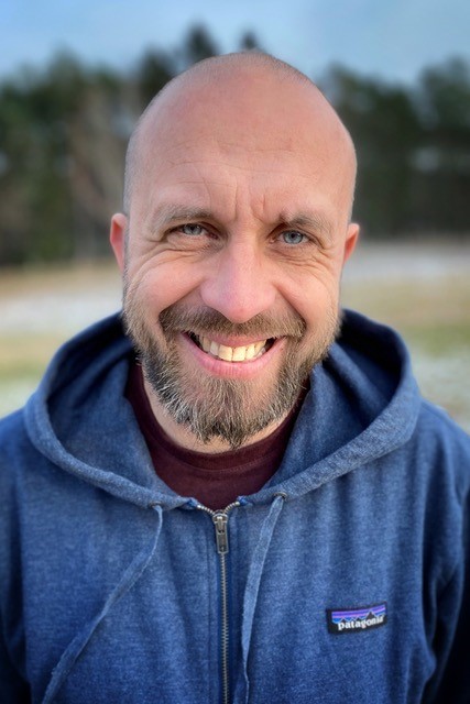 Zdjęcie Roberta Maciąga. Stoi uśmiechnięty. Głowa łysa, niebieska bluza z opuszczonym na ramiona kapturem.