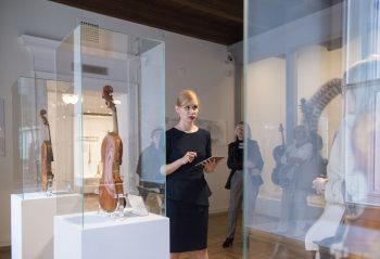 Zdjęcie Pani oprowadzającej po wystawie w Muzeum Instrumentów Muzycznych. Widać szklane gabloty z instrumentami umieszczonymi wewnątrz.