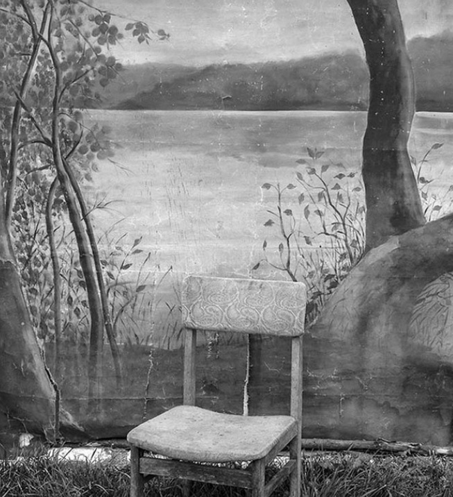 Czarno-biała fotografia przedstawia krzesło stojące na ziemi porośniętej trawą