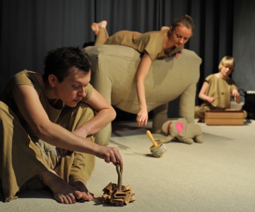 Zdjęcie aktorów w trakcie spektaklu. Trzech aktorów ubranych na beżowo z młynkowymi instrumentami. Beżowy słonik z różowymi uszami.