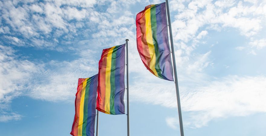 Zdjęcie 3 flag LGBT na masztach na tle niebieskiego nieba z białymi chmurkami.