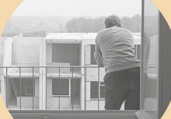 fotografia przedstawia, mężczyznę odwróconego plecami, opierającego się o barierkę balkonu, w tle widać niedokończony wielopiętrowego budynek