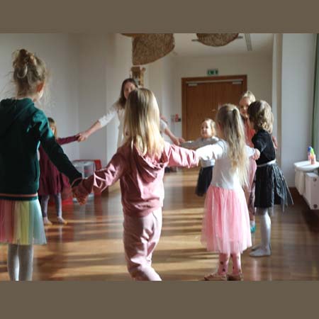 Na zdjęciu grupa dzieci wraz z prowadzącą zajęcia tańczy trzymając się za ręce w kole.
