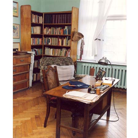 Wnętrze mieszkania-pracowni poetki. Na pierwszym planie stół zastawiony dokumentami, w tle regał z książkami.