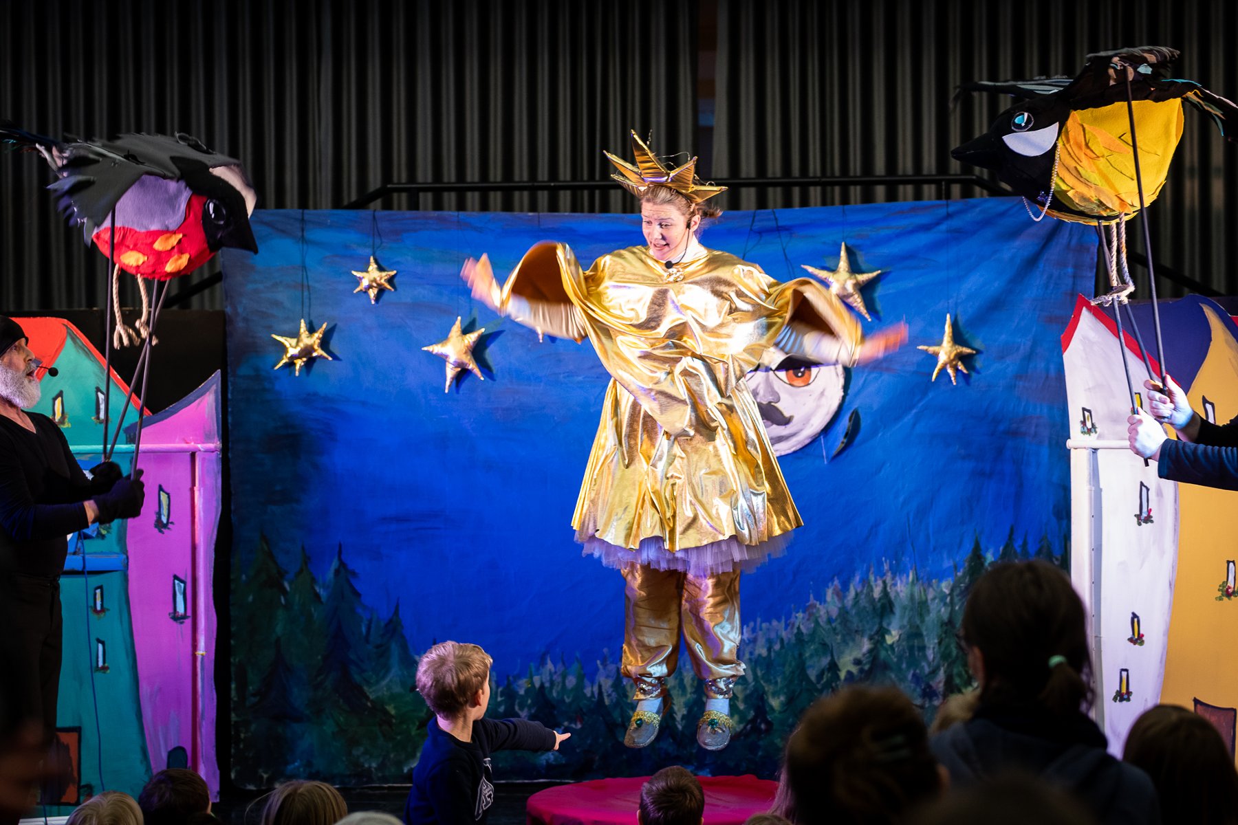 Kadr ze spektaklu. Kobieta ubrana w złoty strój gwiazdki wisi zawieszona na tle niebieskiej płachty symbolizującej niebo. Przed sceną widać chłopca i głopwy kilku innych osób.