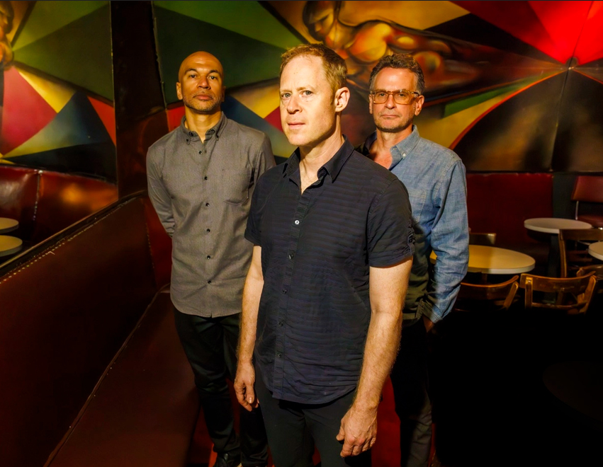 Członkowie zespołu pozują do zdjęcia w zaciemnionych wnętrzach klubu. Za nimi bordowa kanapa narożnikowa i stolik.