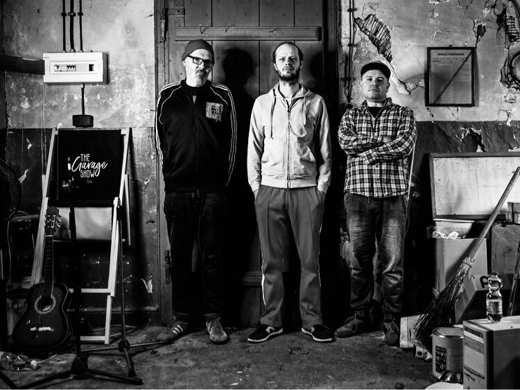 Czarno-białe zdjęcie, na którym widać członków tria pozujących na tle drzwi.