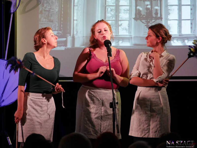 Zdjęcie ze spektaklu. Trzy aktorki w trakcie występu stoją przy mikrofonie.