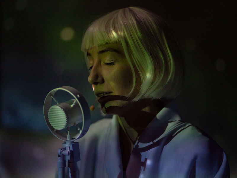 na zdjęciu twarz kobiety o jasnych włosach, z grzywką, przed nią mikrofon. Kobieta ma zamknięte oczy.