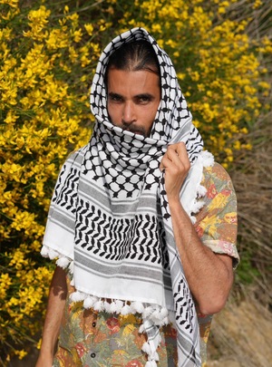 Grafika na tle miętowego koloru. Na środku znajduje się zdjęcie mężczyzny z brodą i długimi ciemnymi włosami, ubranego w kefiję.