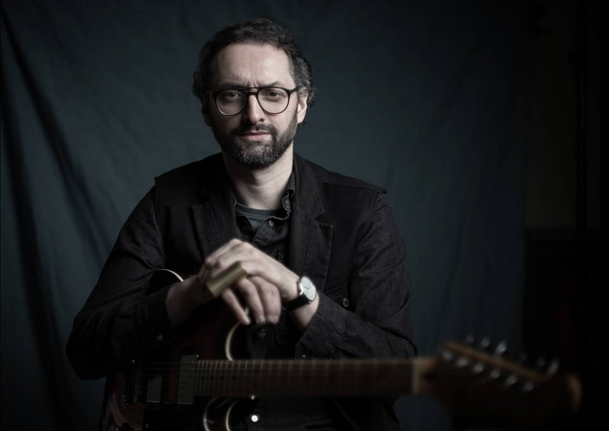 Mężczyzna w ciemnym stroju, trzymający skrzyżowane ręce na gitarze przed sobą. Ciemne tło.