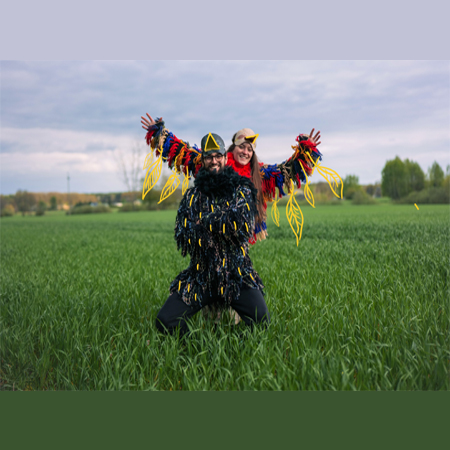 Kobieta i mężczyzna przebrani za kolorowe ptaki pozują do zdęcia. Stoją na zielonej polanie.