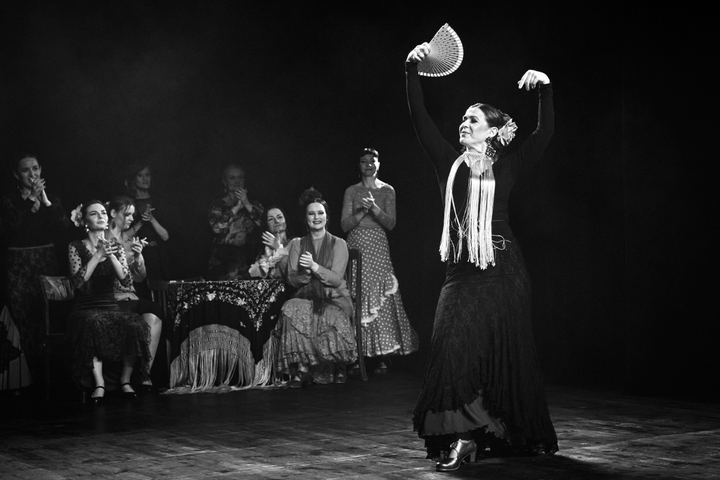 Czarno-białe zdjęcie tancerki flamenco w tanecznej pozie z wachlarzem. Na drugim placnie kilka kobiet obserwujących występ i klaszczących w dłonie.