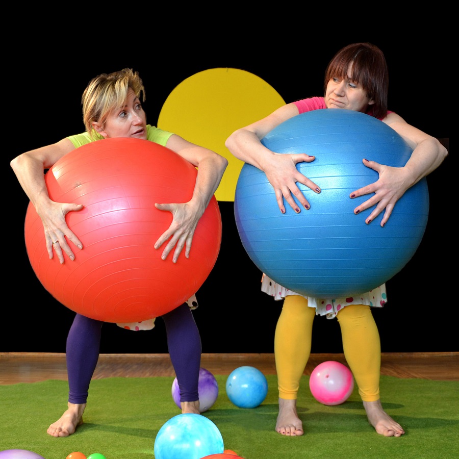 Dwie kolorowo ubrane kobiety, stojące boso na scenie, jedna z nich trzyma przed sobą dużą czerwoną piłkę, druga niebieską.